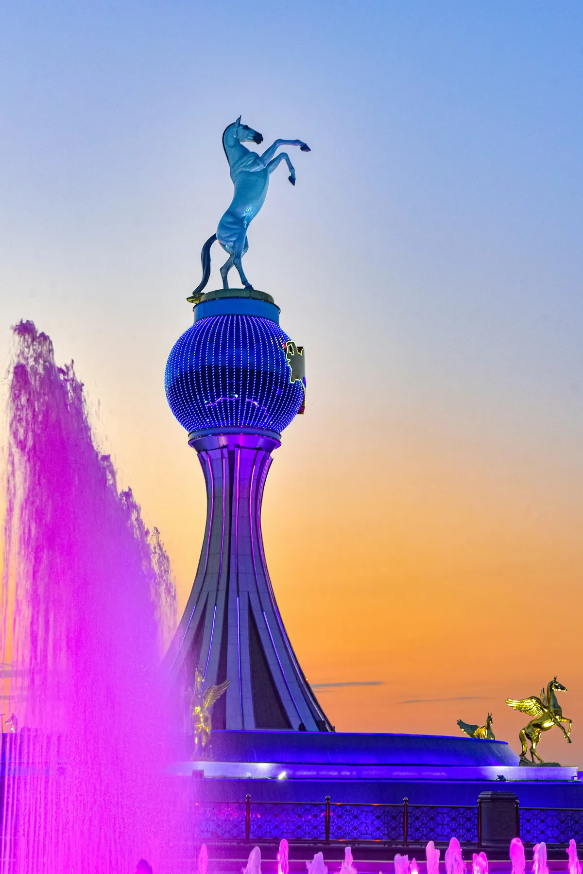 Statue of an Akhal-Teke horse on a blue glowing sphere, at the base of the column are golden winged horses and a fountain with bright pink lighting against the evening sky in Ashgabat
