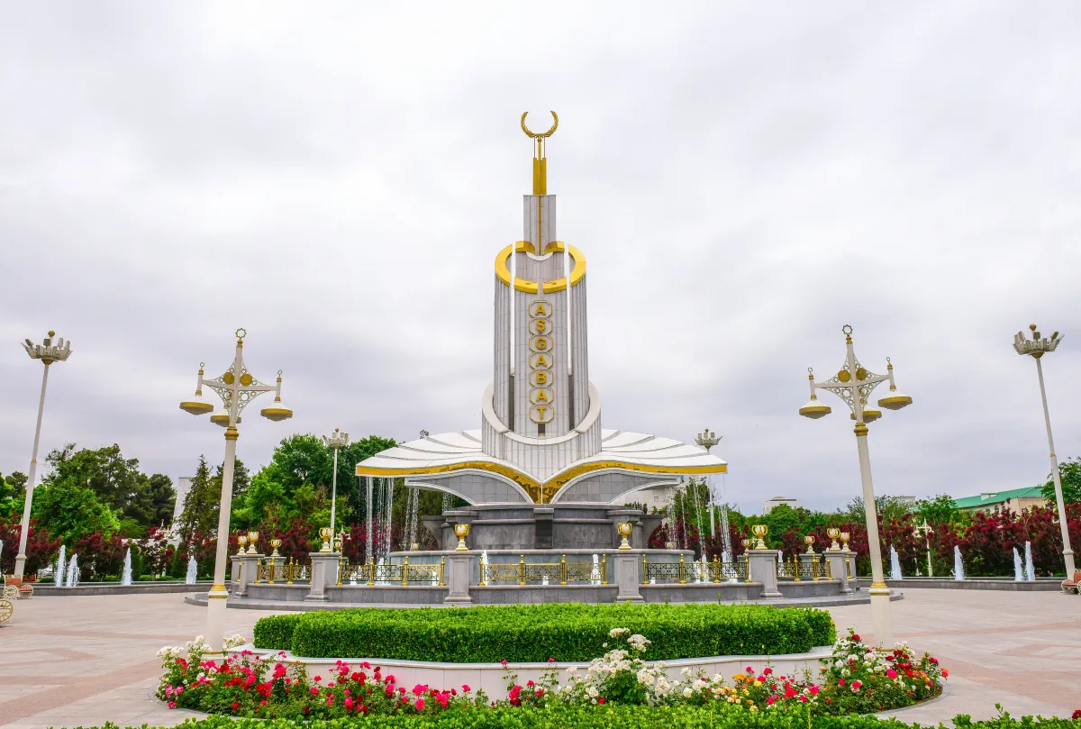 White-marble monument with a golden vertical inscription 'Ashgabat' and fountains, surrounded by blooming roses in a park