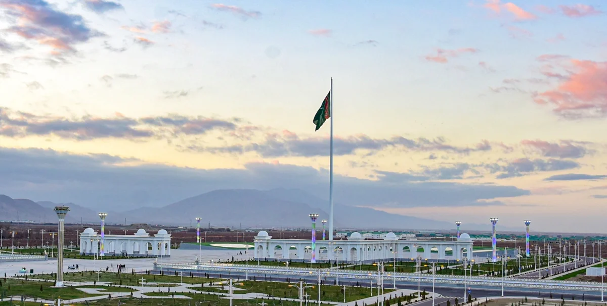 Giant Turkmenistan flag on a tall flagpole at a city square with mountains in the background at sunset