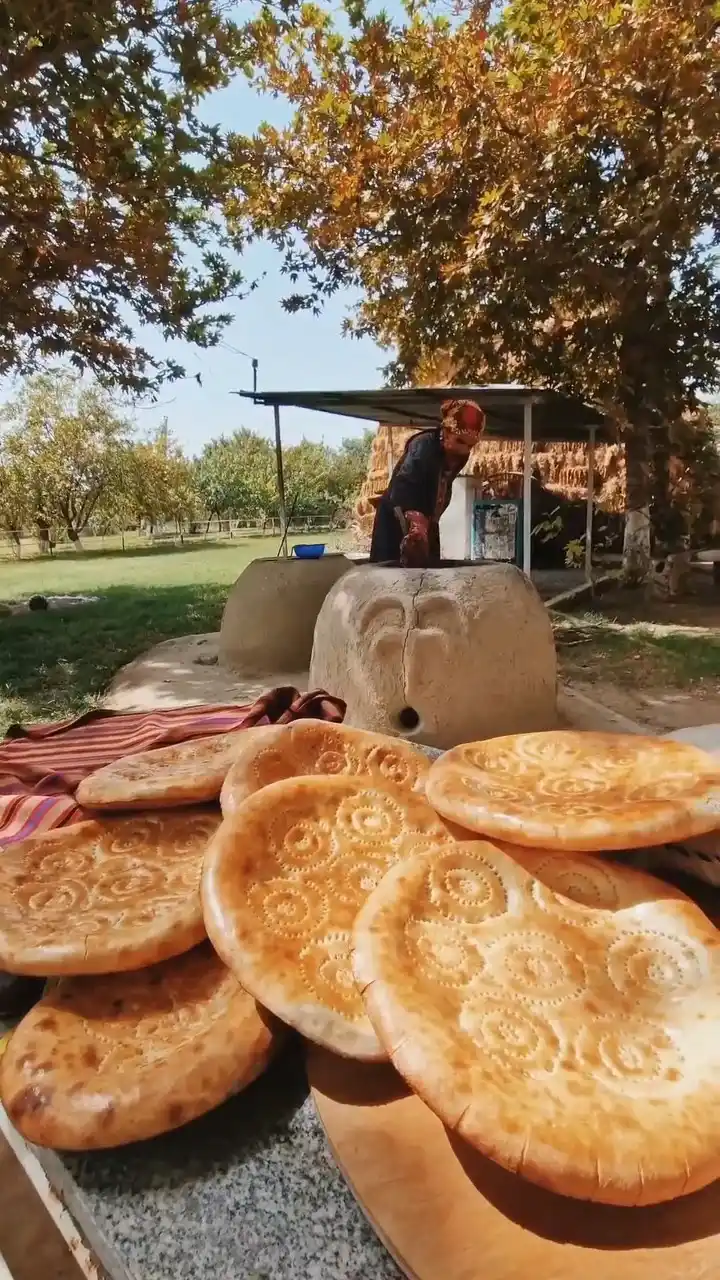 Turkmen woman baking traditional bread in a clay tandoor; hot patterned round loaves in the foreground—authentic village experience in Turkmenistan.